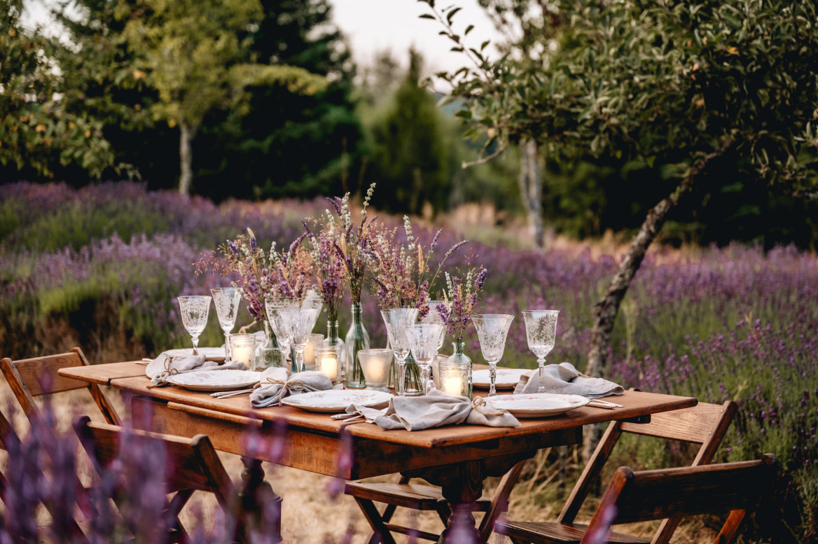 A 6 person table set up in a field of lavendar. A wooden table, 6 chairs, vases of purple, white and blush coloured florals, vintage white crockery with pink floral patterns, clear glassware and napkins tied with string.