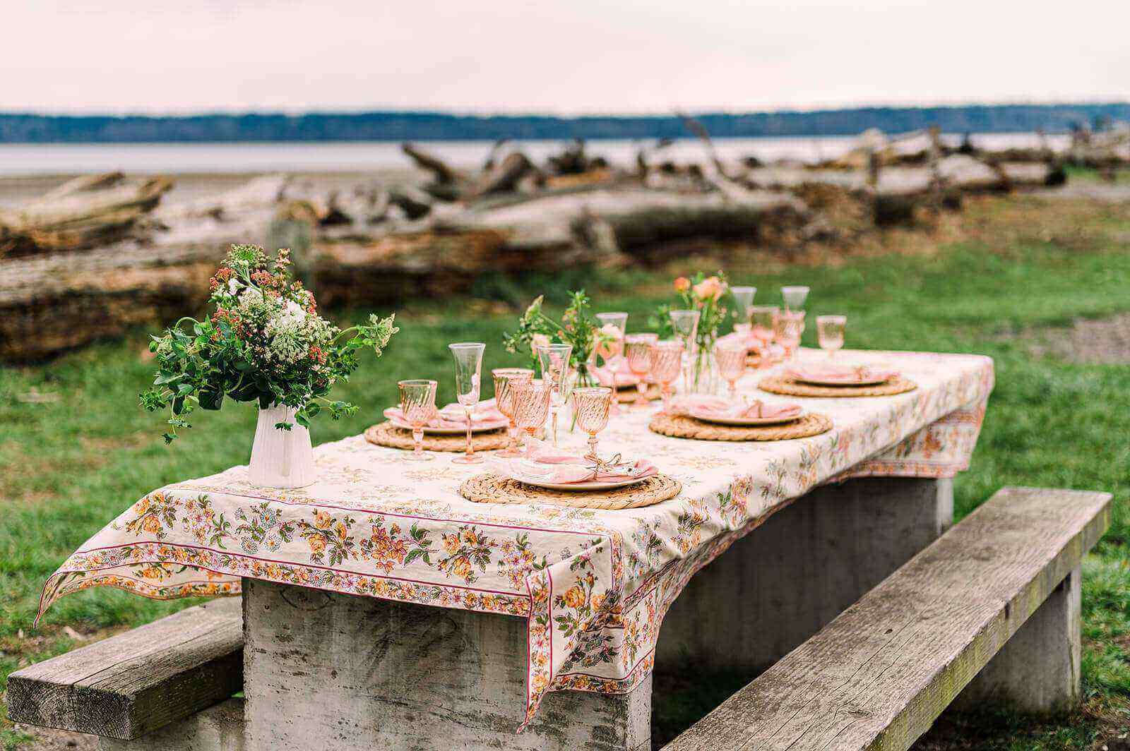 Beachside picnic set up. Wooden picnic bench seating, blush glassware, flowers in a white jug, wicker placemats, pink napkins tied with string, patterned crockery and a floral patterned table cloth