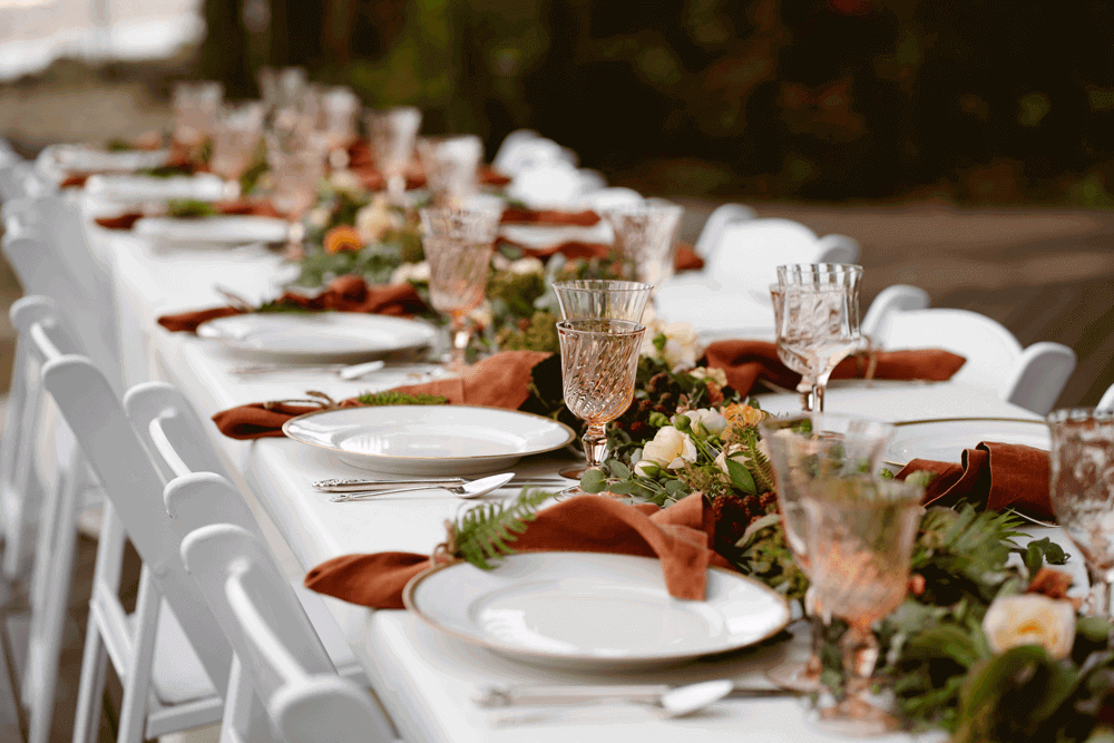 Fresh fern table runner along the middle of the entire table. Cinnamon coloured napkins tied with string and fern. Blush glassware, white table and chairs, silver cutlery and white crockery with gold edging.