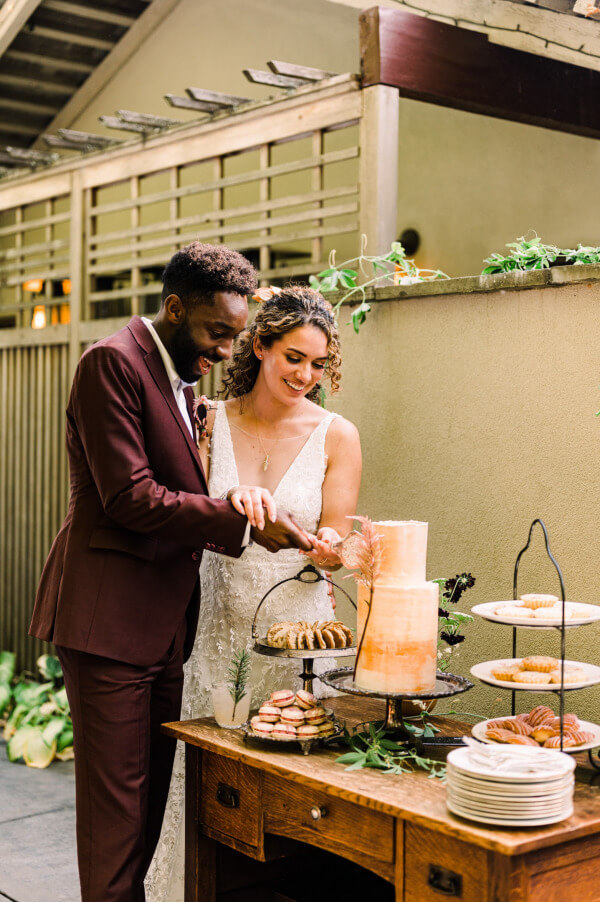 A bride in a white dress and a groom in a maroon suit, cutting their wedding cake, which is surrounded by two multi-layered cake stands filled with desserts, all foods are displayed on a wooden table.