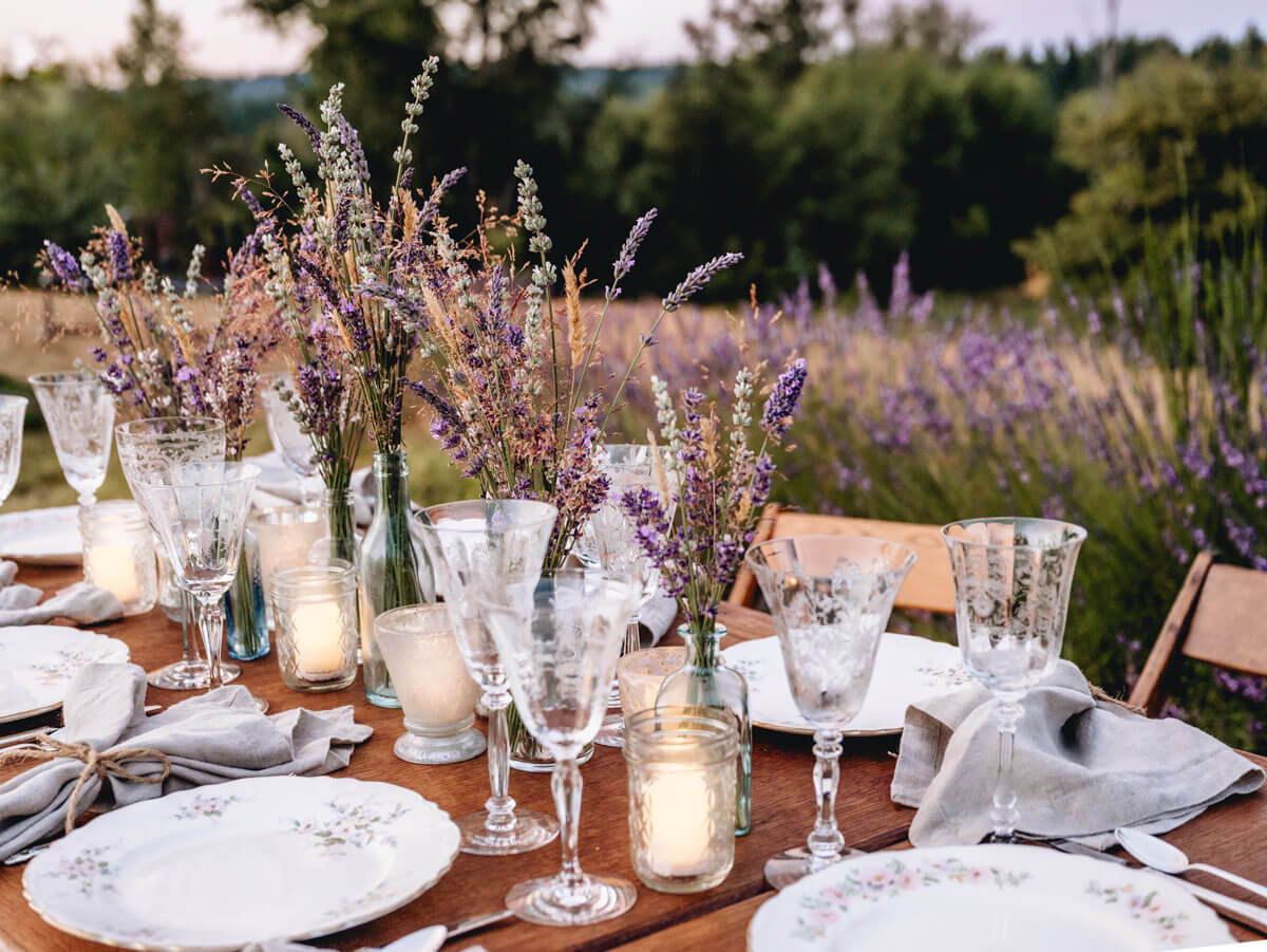 A table set up in a field of lavendar. A wooden table, 6 chairs, vases of purple, white and blush coloured florals, vintage white crockery with pink floral patterns, clear glassware and napkins tied with string.