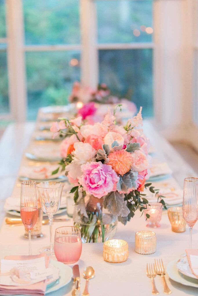 A table set up with blush and clear glassware, white crockery, gold cutlery and gold reflective tealight holders. Pink, orange, white and green floral arrangements as centrepieces. A blurred white paned window in the background.