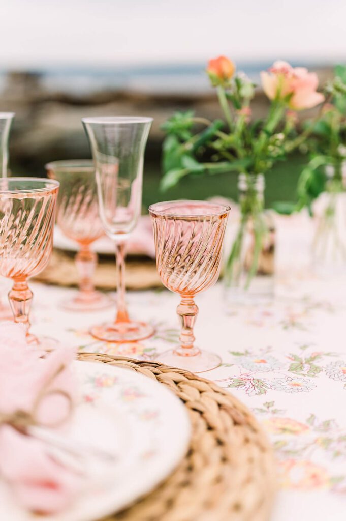 Blush pink glassware in focus, blush flowers in glass vases blurred in the background. A wicker charger plate, white crockery with pink flowers, pink napkin tied with string blurred in the foreground.