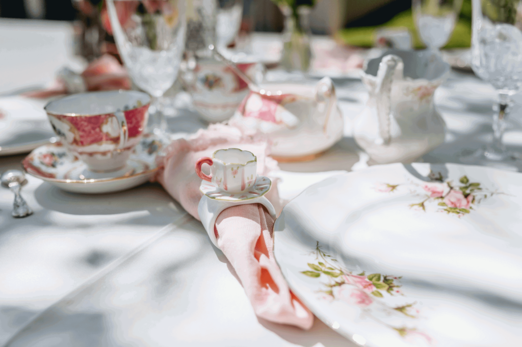 White, pink and gold colour theme. Vintage plates, teacups and saucers, pink napkins, napkin rings with a 3D teacup on them. Clear glassware and silver cutlery on a white tablecloth.