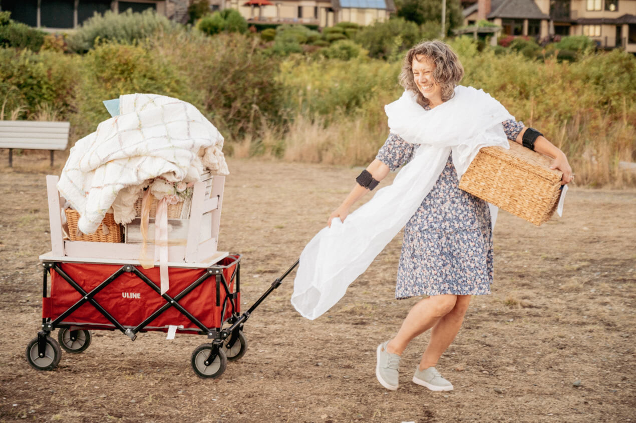 Smiling woman is pulling a red wagon piled high with quilts through a field. There are houses in the distance and a park bench in front of the hedges in the background.
