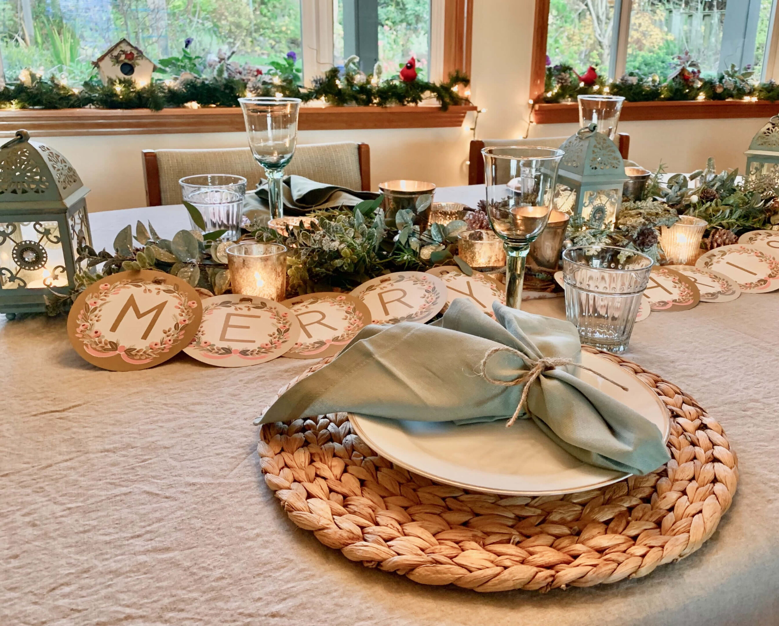 Soft sage green Christmas table set up. Muted green glassware, sage napkins tied with string, sage coloured lanterns, wicker place mats, with a gold and blush coloured 'Merry Christmas' sign, candles and greenery for the table decor.