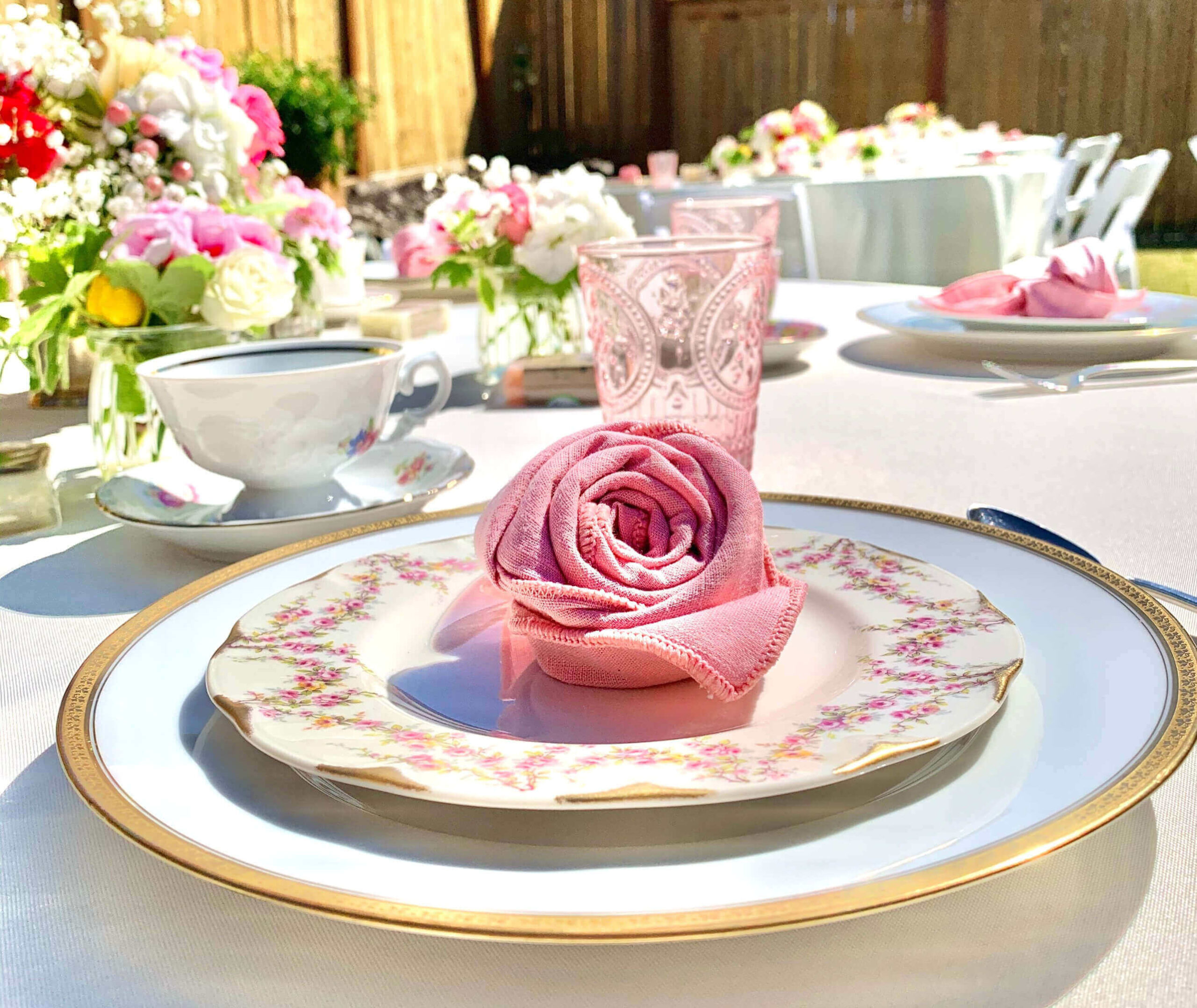 A close up on a tabe set up, pink napkin shaped into a rose bud, vintage crockery with pink floral patterns, a white charger plate with gold edging. Pink glassware, vintage crockery tea sets and bright florals