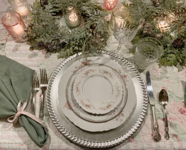 A soft sage green, blush pink and white floral tablecloth, silver charger plate, silver cutler, white crockery with pink and soft sage green floral pattern, sage green napkin tied with a pink bow, crystal glassware, pink glass tealightholders with lit teakights inside and leafy wreaths for decoration on the table.