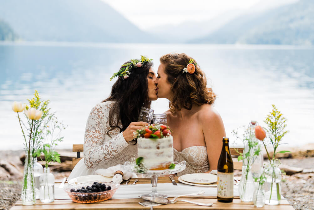 Two brides are sitting at a picnic table kissing, sat at a beautifully decorated table including a naked wedding cake, flowers in vases and fruit. They are sat in front of a hilly landscape and a lake.