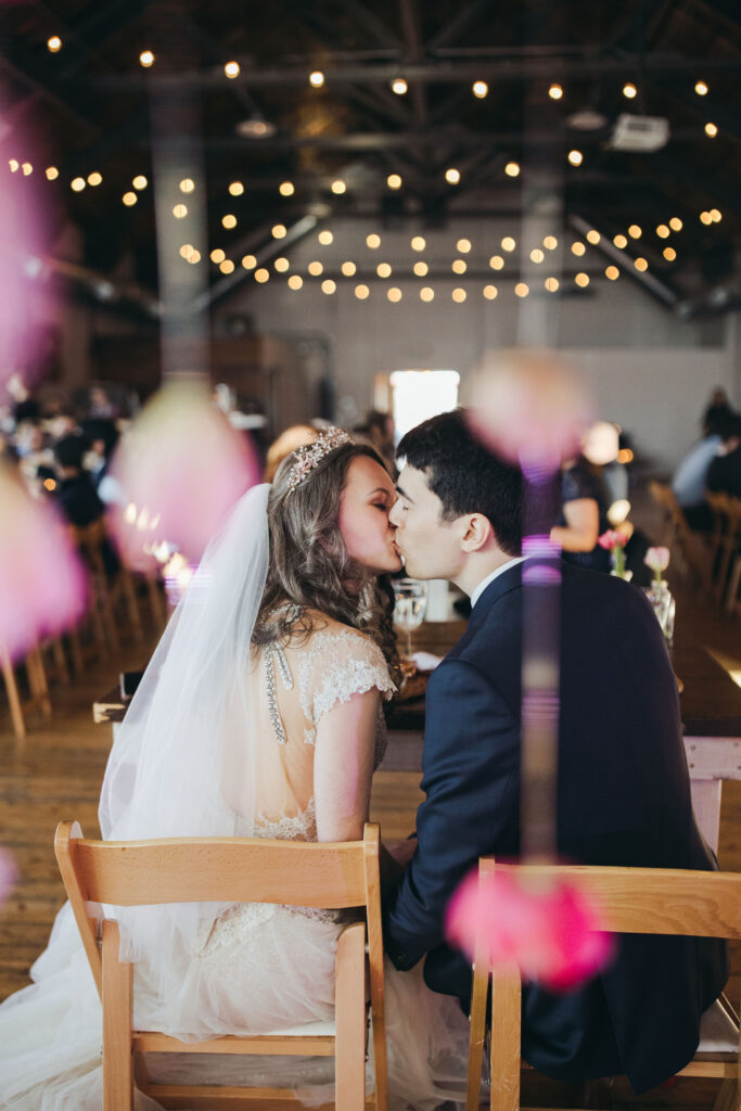 Bride and Groom share a kiss at their sweetheart table with their guests at tables in front of them. Photo taken from behing the newlyweds.