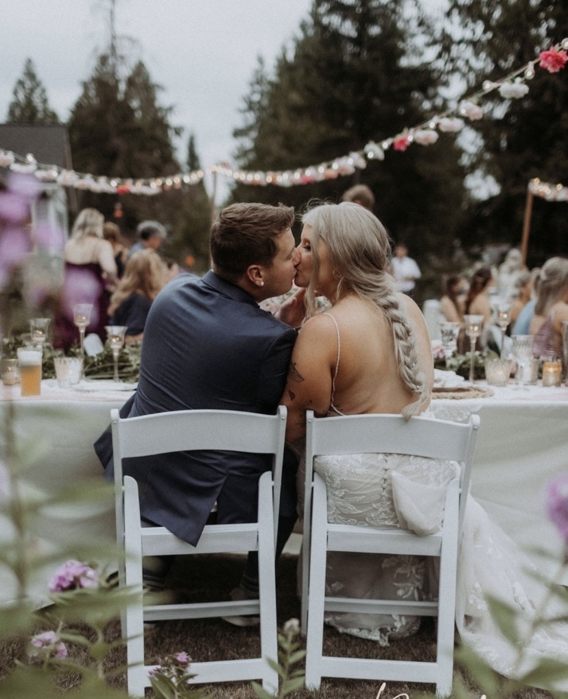Bride and Groom share a kiss at their sweetheart table with their guests at tables in front of them at their outdoor wedding set up. Photo taken from behing the newlyweds.