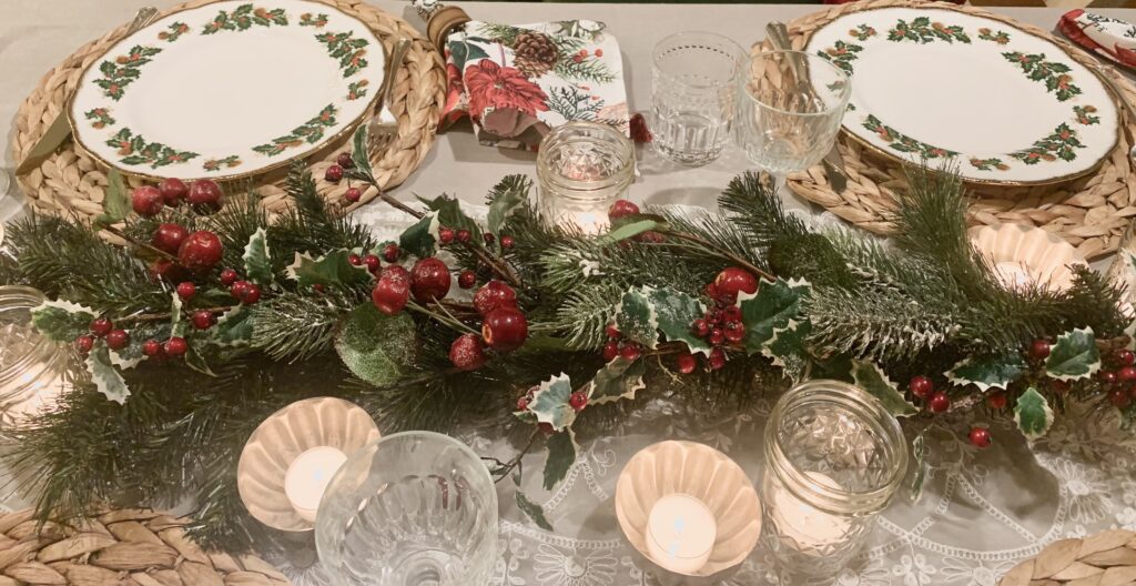 Christmas table setting with faux evergreen garland that has holly, white tipped fir branches, and red berries, surrounded by glowing gold votive candles, and set with white dishes that have a holly border around them.