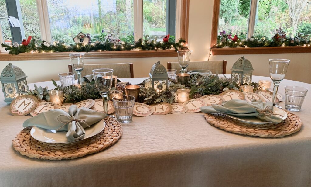 Christmas table setting with a light colored tablecloth, woven placemats, and soft sage green napkins with sparkly soft evergreen garland and candles, and floral letters that spell out Merry Christmas.